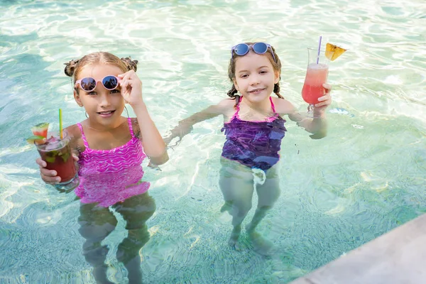 Ragazze in costume da bagno con cocktail di frutta guardando la fotocamera mentre in piedi in piscina — Foto stock