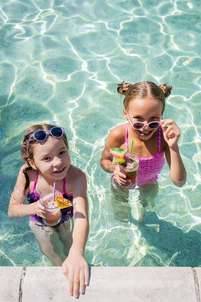 Vista de alto ângulo da menina tocando óculos de sol enquanto estava na piscina perto de amigo com cocktail refrescante — Fotografia de Stock