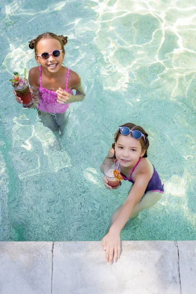 Высокий угол обзора девушек, смотрящих в камеру, держа стаканы с фруктовыми коктейлями в бассейне — стоковое фото