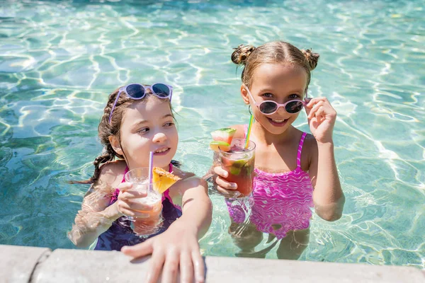 Foco seletivo de meninas em fatos de banho e óculos de sol segurando coquetéis de frutas frescas na piscina — Fotografia de Stock