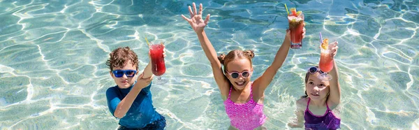 Cultivo panorámico de niñas y niños de pie en la piscina y la celebración de cócteles refrescantes en las manos levantadas - foto de stock