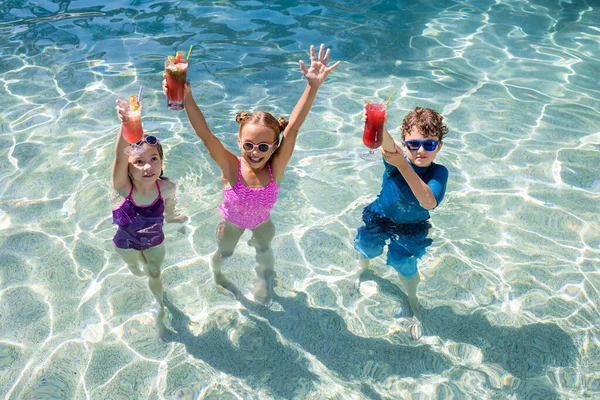 Высокий угол обзора детей, стоящих в бассейне и держащих свежие фруктовые коктейли в поднятых руках — стоковое фото