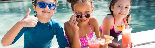 Horizontal crop of boy showing thumb up near girls with refreshing cocktails at poolside — Stock Photo