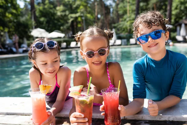 Amigos alegres en gafas de sol y trajes de baño celebración de cócteles de frutas frescas cerca de la piscina — Stock Photo