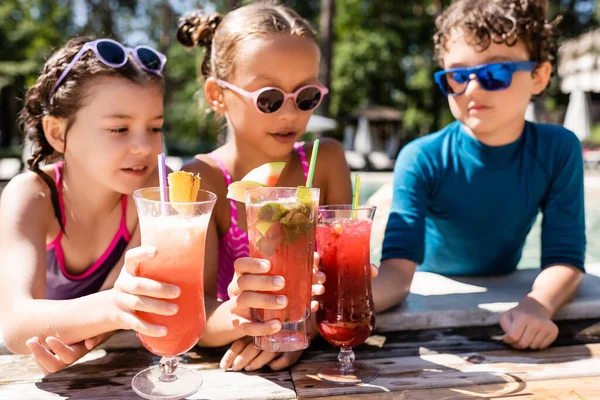 Fuoco selettivo di amici che si accarezzano occhiali con cocktail di frutta fresca vicino a piscina — Foto stock