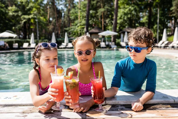 Amigos en trajes de baño tintineo vasos con cócteles de frutas frescas cerca de la piscina — Stock Photo