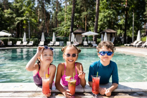 Menina em óculos de sol mostrando gesto de vitória perto de amigos com coquetéis de frutas frescas na beira da piscina — Fotografia de Stock