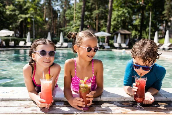 Chico en gafas de sol beber cóctel de fruta fresca cerca de las niñas en trajes de baño en la piscina - foto de stock