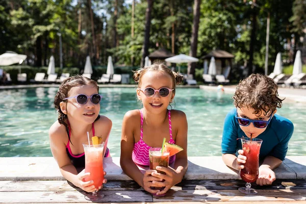 Joyful friends in swimwear and sunglasses with fresh fruit cocktails at poolside — Stock Photo