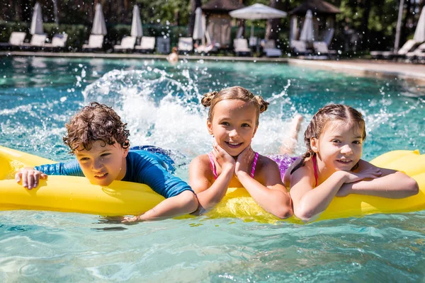 Amigos alegres que fazem respingos de água com pernas enquanto flutuam na piscina no colchão inflável — Fotografia de Stock