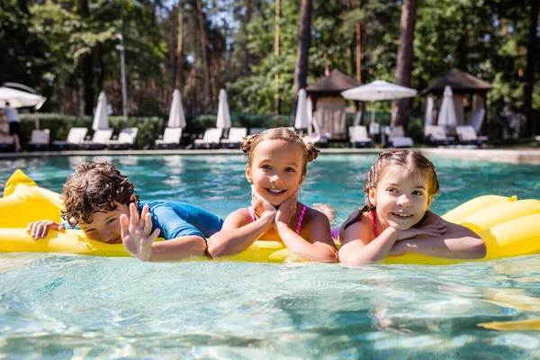 Joyful boy waving hand at camera while swimming on inflatable mattress with girls — Stock Photo