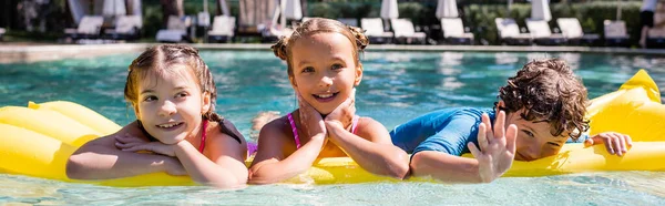 Panoramic concept of boy waving hand at camera while floating on inflatable mattress with girls — Stock Photo