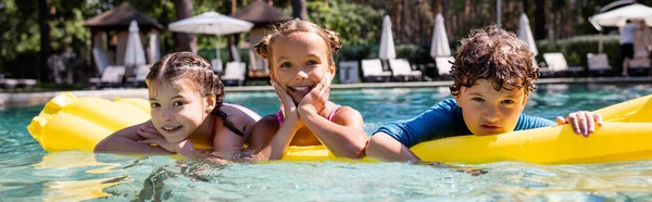 Imagem horizontal de amigos alegres que olham para a câmera enquanto flutuam no colchão inflável na piscina — Fotografia de Stock