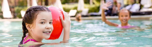 Enfoque selectivo de la muchacha que sostiene la bola inflable cerca de amigo que agita la mano en la piscina, concepto panorámico - foto de stock