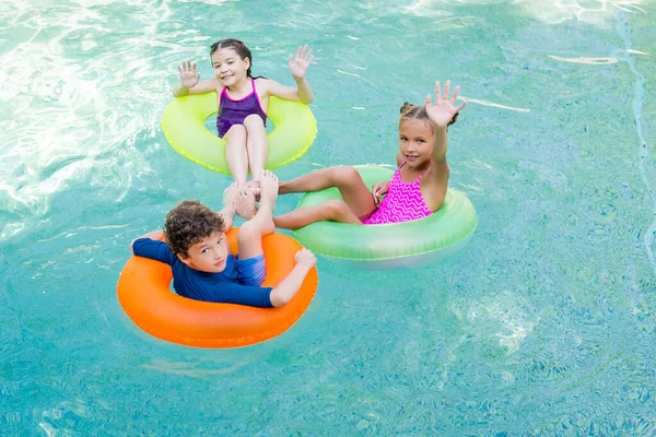 Vue grand angle d'amis joyeux agitant les mains tout en nageant dans la piscine sur des anneaux gonflables — Photo de stock