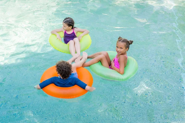 Vue grand angle de fille regardant la caméra tout en flottant dans la piscine sur des anneaux gonflables avec des amis — Photo de stock