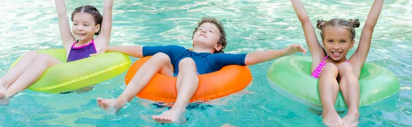 Panoramic concept of girls with hands in air and boy with closed eyes swimming in pool on inflatable rings — Stock Photo