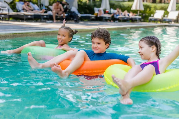 Довольный мальчик и две девочки плавают в бассейне на красочных надувных кольцах — стоковое фото