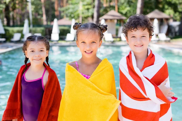 Amigos mojados mirando a la cámara mientras envuelven en toallas de felpa cerca de la piscina — Stock Photo