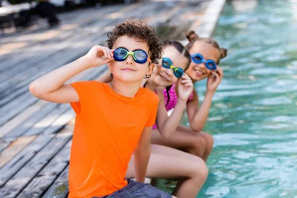 Foco seletivo de amigos tocando óculos de natação enquanto sentado perto da piscina e olhando para a câmera — Fotografia de Stock