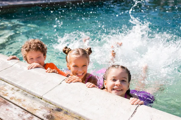 Amici eccitati guardando la fotocamera mentre fanno spruzzi d'acqua con le gambe in piscina — Foto stock