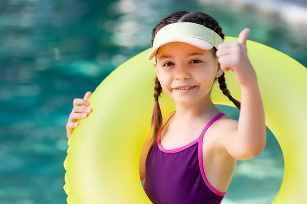 Chica en traje de baño y gorra del visor de sol que muestra el pulgar hacia arriba mientras sostiene el anillo inflable - foto de stock