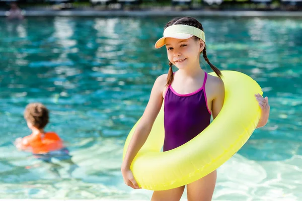 Chica en traje de baño y gorra de visera de sol posando con anillo inflable cerca de la piscina - foto de stock