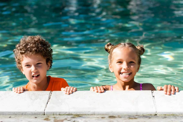 Aufgeregtes Mädchen blickt in die Kamera, während es Zeit im Pool neben lockigem Jungen verbringt — Stockfoto