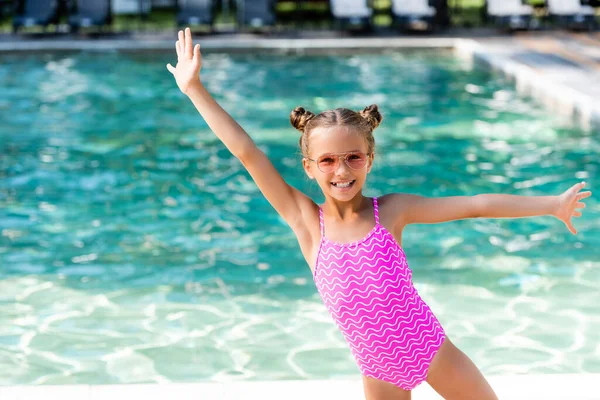 Menina em roupa de banho e óculos de sol de pé com as mãos estendidas perto da piscina — Fotografia de Stock