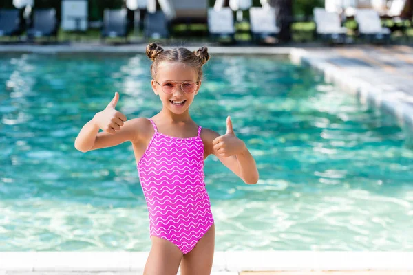 Chica en traje de baño y gafas de sol mostrando los pulgares hacia arriba mientras está de pie cerca de la piscina - foto de stock