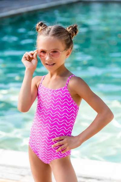 Niño en traje de baño tocando gafas de sol mientras posa con la mano en la cadera cerca de la piscina - foto de stock