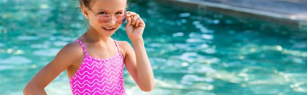 Горизонтальная концепция девушки в купальнике, касающейся солнцезащитных очков при взгляде на камеру возле бассейна — стоковое фото