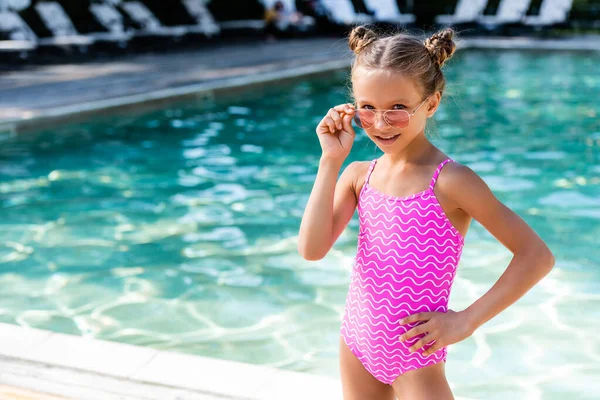 Mädchen im Badeanzug berührt Sonnenbrille, während sie mit der Hand auf der Hüfte am Pool steht — Stockfoto