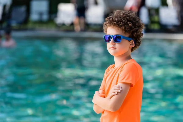 Enfant bouclé en lunettes de soleil bleues et t-shirt orange debout avec bras croisés près de la piscine — Photo de stock