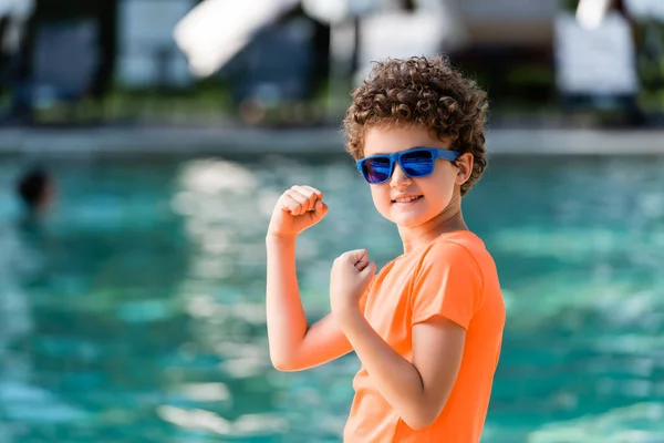 Niño rizado en camiseta naranja y gafas de sol demostrando fuerza mientras mira a la cámara - foto de stock