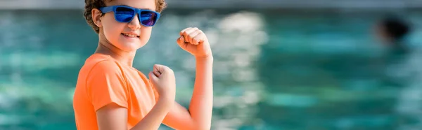 Plan panoramique de garçon en t-shirt orange et lunettes de soleil démontrant la puissance — Photo de stock