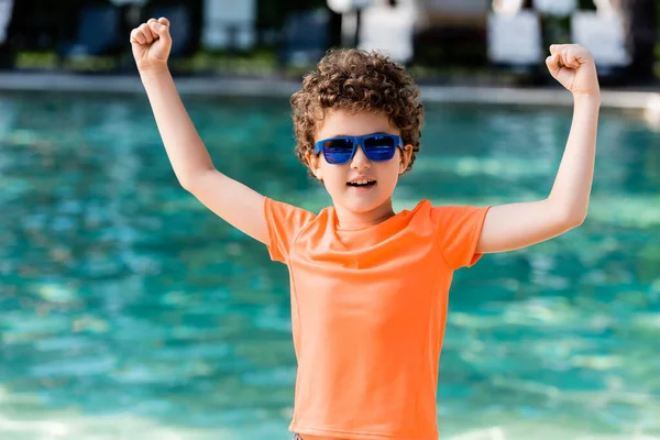 Niño rizado en camiseta naranja y gafas de sol mostrando el gesto ganador cerca de la piscina - foto de stock