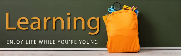 Horizontal image of yellow backpack full of school stationery near green chalkboard with learning, enjoy life while you are young lettering — Stock Photo