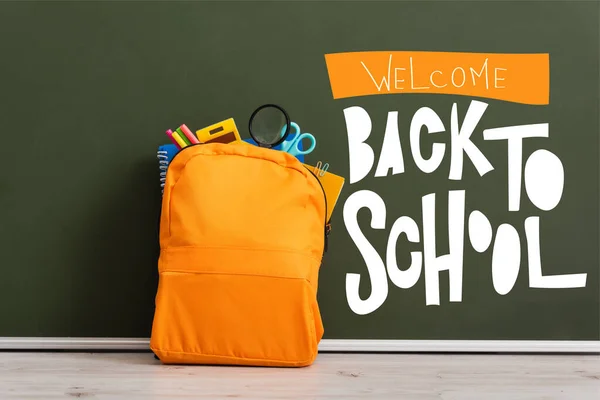 Mochila amarilla llena de papelería escolar cerca de pizarra verde con bienvenida de nuevo a las letras de la escuela - foto de stock