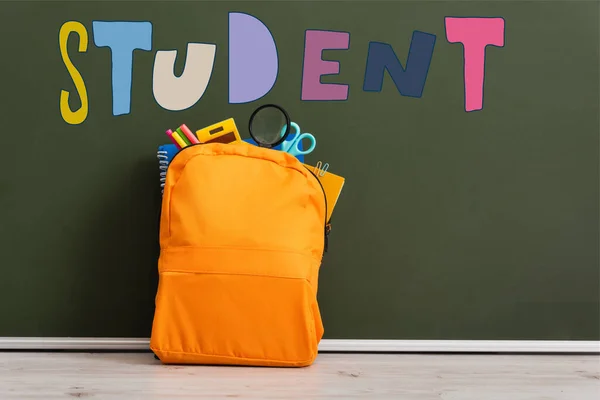 Mochila amarilla llena de papelería escolar cerca de pizarra verde con letras de estudiante - foto de stock