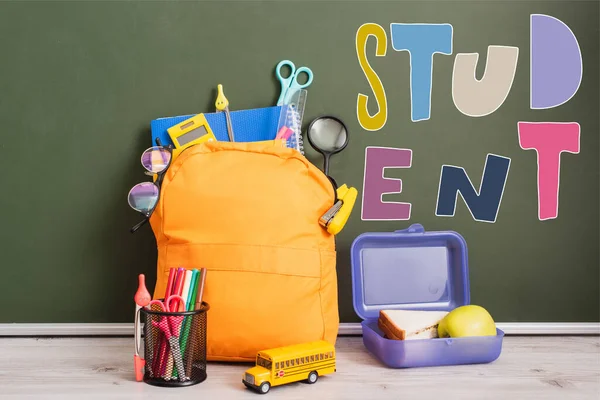 Gelber Schulrucksack voller Schulbedarf in der Nähe der Brotdose, Spielzeugschulbus und Stifthalter auf dem Schreibtisch neben grüner Tafel mit Schülerschriftzug — Stockfoto