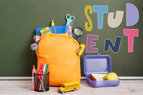 Mochila amarela cheia de material escolar perto da lancheira, ônibus escolar de brinquedo e suporte de caneta na mesa perto de quadro verde com letras estudantis — Fotografia de Stock