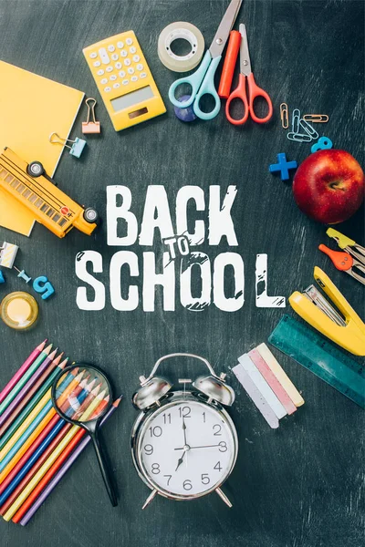 Vista superior del marco con manzana, autobús escolar de juguete, reloj despertador vintage y útiles escolares en pizarra negra con letras de regreso a la escuela - foto de stock