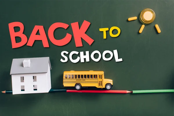 Vista superior del autobús escolar de juguete en la carretera hecha de lápices de color, modelo de casa y sol en pizarra verde con letras de vuelta a la escuela - foto de stock