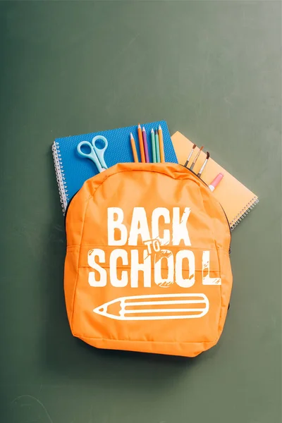 Vista superior de la mochila repleta de papelería escolar en pizarra verde con letras de vuelta a la escuela e ilustración a lápiz - foto de stock