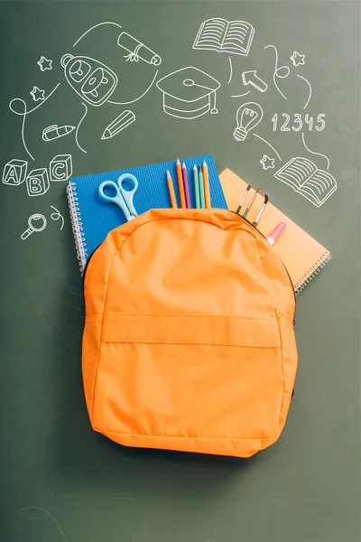 Top view of backpack packed with school stationery on green chalkboard with illustration — Stock Photo