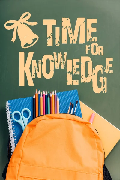 Top view of yellow backpack with notebooks, scissors, paintbrushes and color pencils on green chalkboard with time for knowledge lettering — Stock Photo