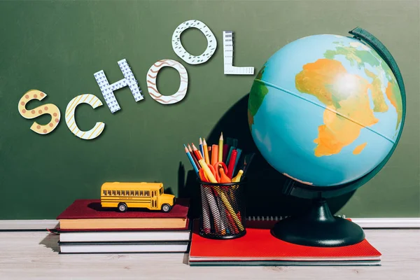 Глобус и держатель ручки на ноутбуке возле игрушечного школьного автобуса на книгах возле зеленой доски со школьными надписями — стоковое фото