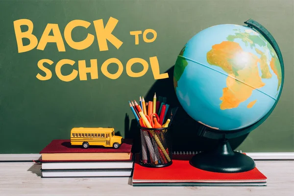 Globus und Stifthalter auf Notizbuch in der Nähe von Spielzeugschulbus auf Büchern in der Nähe von grüner Tafel mit Schulschriftzug — Stockfoto