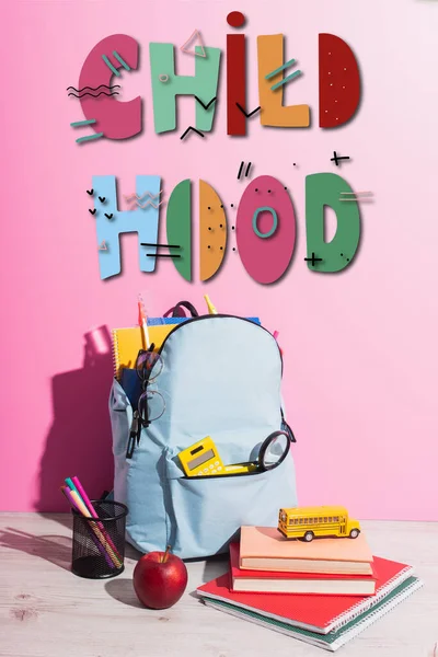 Шкільний рюкзак, повний канцелярських товарів, ручки власника, стигле яблуко, книги та іграшковий шкільний автобус поблизу дитячого садка на рожевому — стокове фото